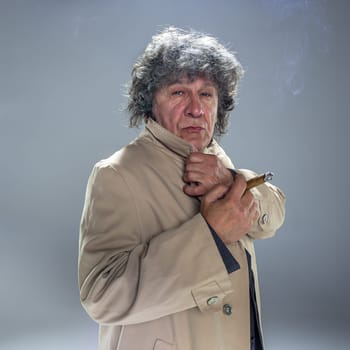 The senior man in cloak with cigar as detective or mafia boss. Studio shot on gray in retro stile