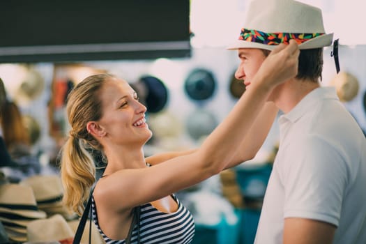 Happy young couple are exploring Queen Victoria Market in Australia. The woman has found a childs summer hat and is trying to squeeze it on her boyfriends head, as he pulls a face.