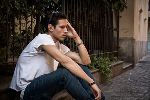 Sad, unhappy young man outdoor, sitting on pavement, holding head with his hand