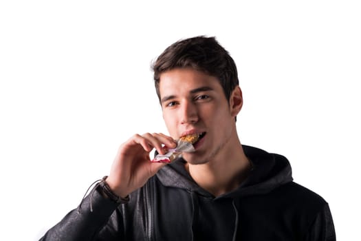 Handsome young man eating cereal bar, looking at camera standing, isolated on white background