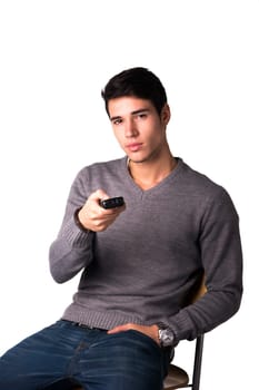 Handsome young man holding remote control and changing TV channels, sitting isolated on white