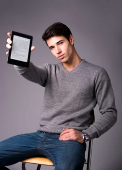 Handsome young man holding and showing ebook reader and looking at camera, sitting on grey background