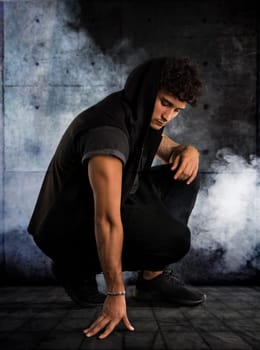 Handsome young man kneeling down in dark hoodie on black background, smoke around him. Looking down