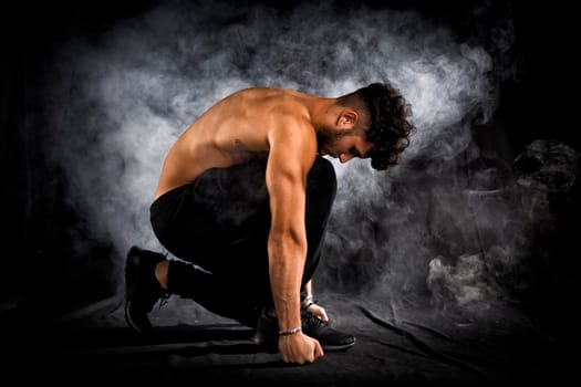 Handsome shirtless muscular young man kneeling down on black background, smoke around him. Looking down