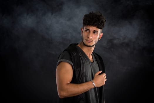 Portrait of handsome young man in dark t-shirt on black background, with smoke around. Looking away