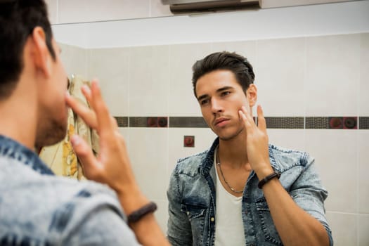 Handsome young man applying moisturizing cream on face in front of home bathroom mirror