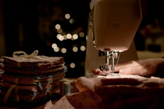 Low key detail of hands working on piece of fabric on automatic sewing machine, shot from the side