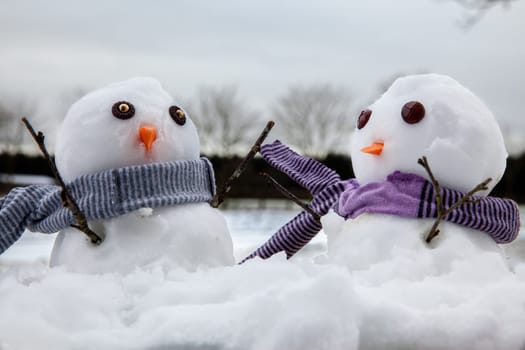 Two cute snowmen wearing scarfs and their twig arms raised in the air. Snow fall all around