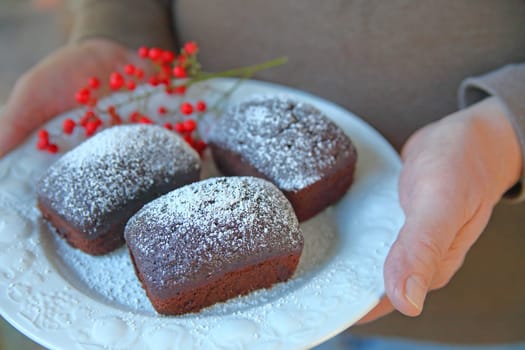 A man holds a decorative plate with three small chocolate cakes dusted with powdered sugar, red berries on the side