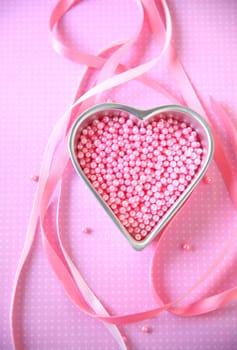 Vertical view of heart-shaped pan of pink cake decoration beads on a polka-dot background with pink ribbons