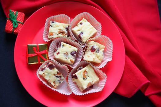 Plate of Christmas fudge made with white chocolate, cranberries and pecans with red napkin and miniature gifts