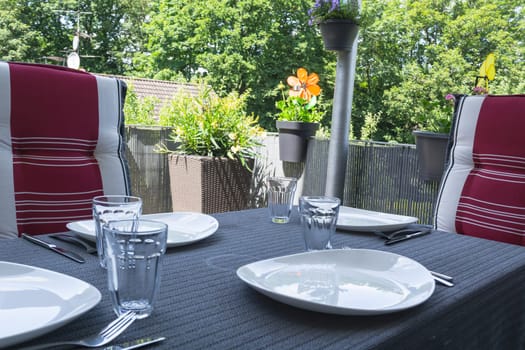 Laid balcony table with white plates,drinking glasses and cutlery with two pieces with modern seat covers.