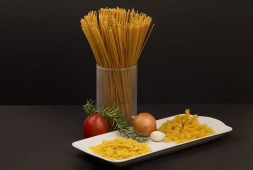 Glass with raw spaghetti and other pasta variants and vegetables on a plate.