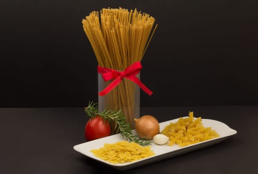 In a glass there are raw spaghetti and on a plate other pasta variants and vegetables.