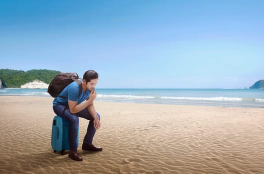 Young asian man with backpack sitting on luggage on the beach
