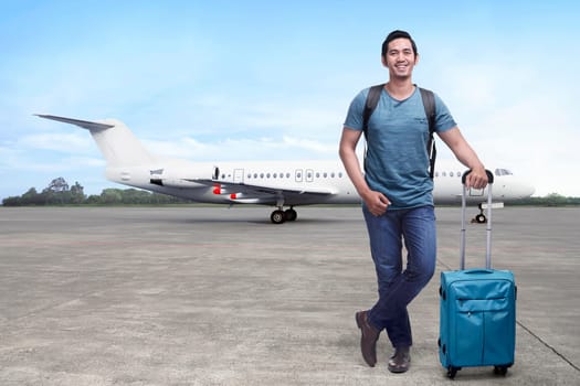 Handsome asian backpacker with suitcase standing against plane background