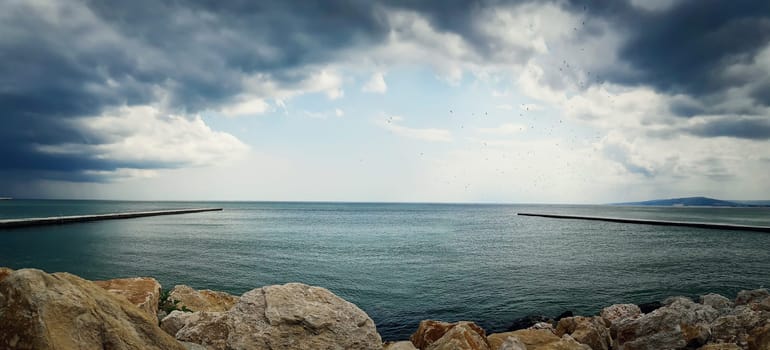 Sea landscape between two piers on the stone beach with a dramatic cloudy sky. Beautiful view at the coast of the Black Sea in Balchik city, Bulgaria.