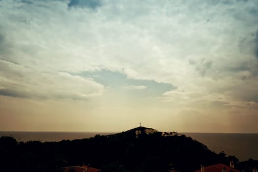 A silhouette of a holy christian cross illuminated by sunlight on top of a hill on a dramatic cloudy sky. Beautiful view at the coast of the Black Sea in Balchik city, Bulgaria.