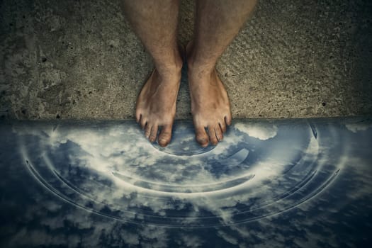 Surrealistic image with male bare legs standing on the edge, in front of water pothole reflecting a cloudy sky.