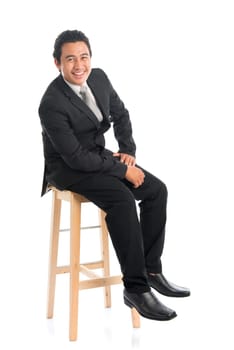 Full body portrait of good looking young Southeast Asian businessman sitting on high chair, isolated on white background.