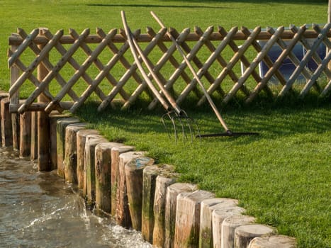 Green lawn of a bathing area on Lake Caldaro with shore fortification,garden fence and garden tools in the evening sun.