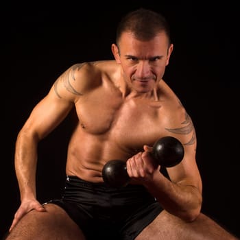 Muscular man exercises his biceps with weight on black background.