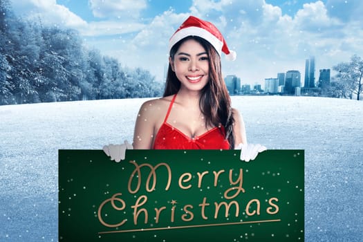 Young attractive asian woman wearing Santa Claus costume holding billboard with merry christmas writing