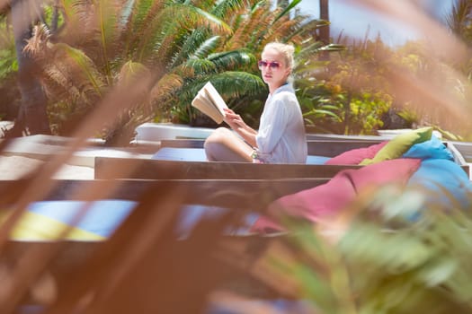 Fancy lady in white beach tunic reading book and relaxing by poolside of luxury spa resort set in lush tropical garden. Relaxation and wellbeing concept. Shot trough palm tree leaves.