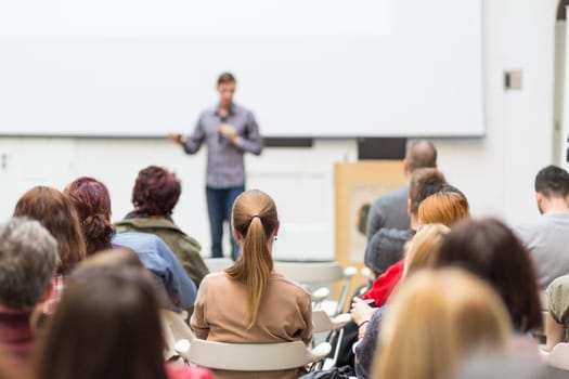 Male speaker giving presentation in lecture hall at university workshop. Audience in conference hall. Rear view of unrecognized participant in audience. Scientific conference event.