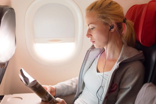 Woman reading magazine and listening to music on airplane. Female traveler reading seated in passanger cabin. Sun shining trough airplane window.