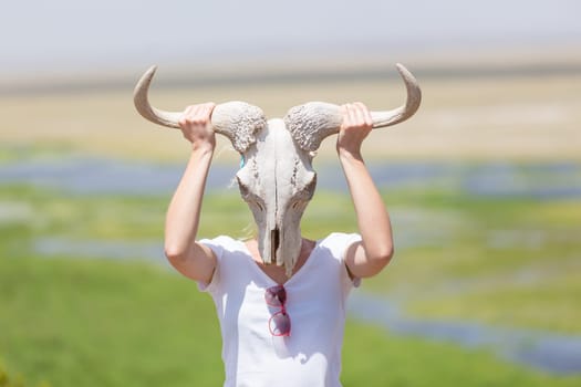 Woman holding a white wildebeest skull wearing it like a mask in nature on african wildlife safari, Amboseli national park, Kenya.