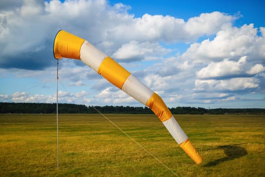 Striped orange and white windsock against blue sky with clouds and field of grass.