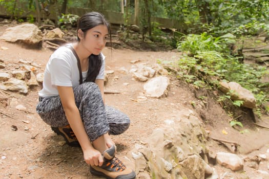 Young asian woman hiker stops to tie her shoe on a summer hiking trail in forest.