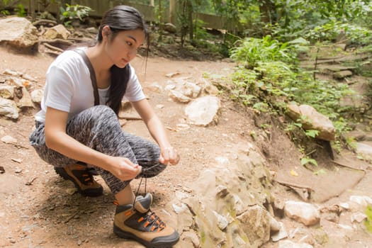 Young asian woman hiker stops to tie her shoe on a summer hiking trail in forest.