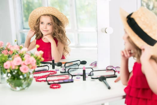 A little girl with red dress and cosmetics. She is in mother's bedroom, sitting near the mirror.