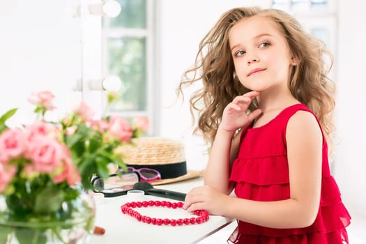 A little girl with red dress and cosmetics. She is in mother's bedroom, sitting near the mirror.