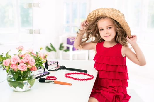 A little girl with red dress and cosmetics. She is in mother's bedroom, sitting near the mirror.