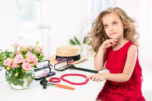 A little girl with red dress and cosmetics. She is in mother's bedroom, sitting near the mirror.
