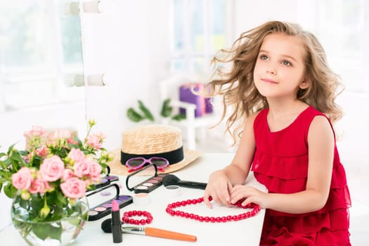 A little girl with red dress and cosmetics. She is in mother's bedroom, sitting near the mirror.