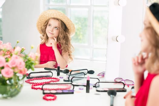 A little girl with red dress and cosmetics. She is in mother's bedroom, sitting near the mirror.