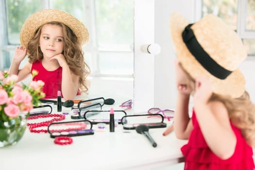 A little girl with red dress and cosmetics. She is in mother's bedroom, sitting near the mirror.