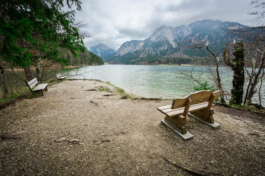 Wide-angled view of the Alpsee with three benches in the foreground on a small rest area near Neuschwanstein Castle in cloudy skies.