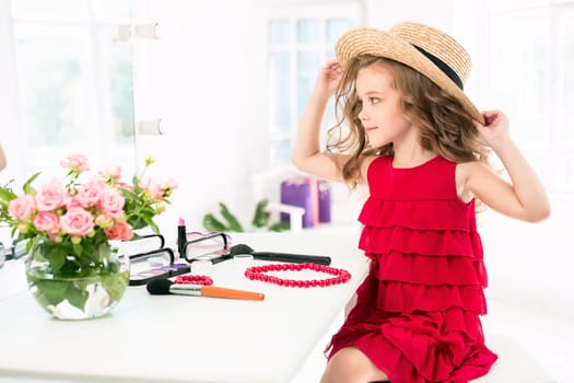 A little girl with red dress and cosmetics. She is in mother's bedroom, sitting near the mirror.