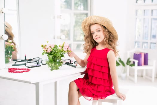 A little girl with red dress and cosmetics. She is in mother's bedroom, sitting near the mirror.