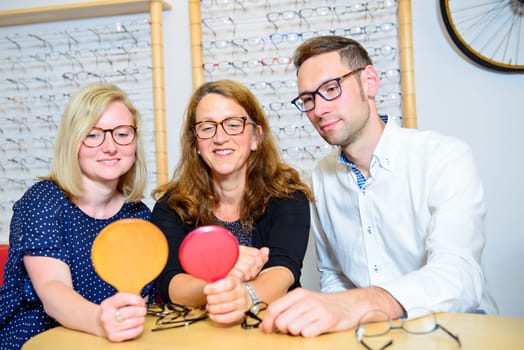 in optician shop-two women and one man with hand mirror up selecting new glasses