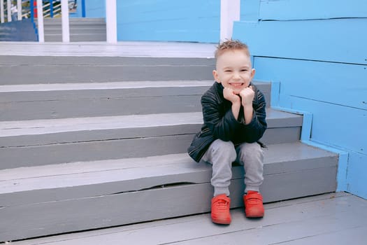 A boy of five, dressed in a leather jacket, in red krosovkah, sitting on the steps, smiling happy life, posing on the camera