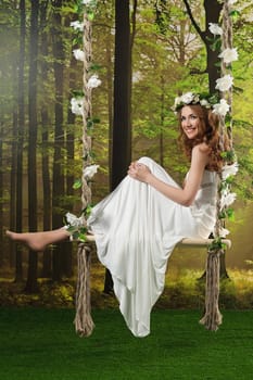Young girl in a long white dress, with a wreath on a swing in the studio.