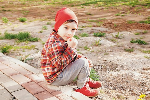 A little boy wearing a red hat, a jacket, and sneakers is sitting in the park. sunny day.