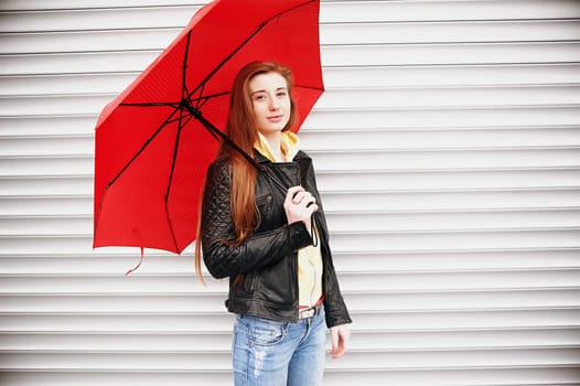 A pretty girl with long red hair, in a black leather jacket with a red umbrella in her hands, walks and smiles. Portrait in the open air