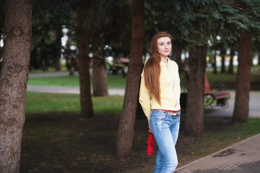 A pretty girl with long red hair, walks through the park and smiles. Portrait in the open air